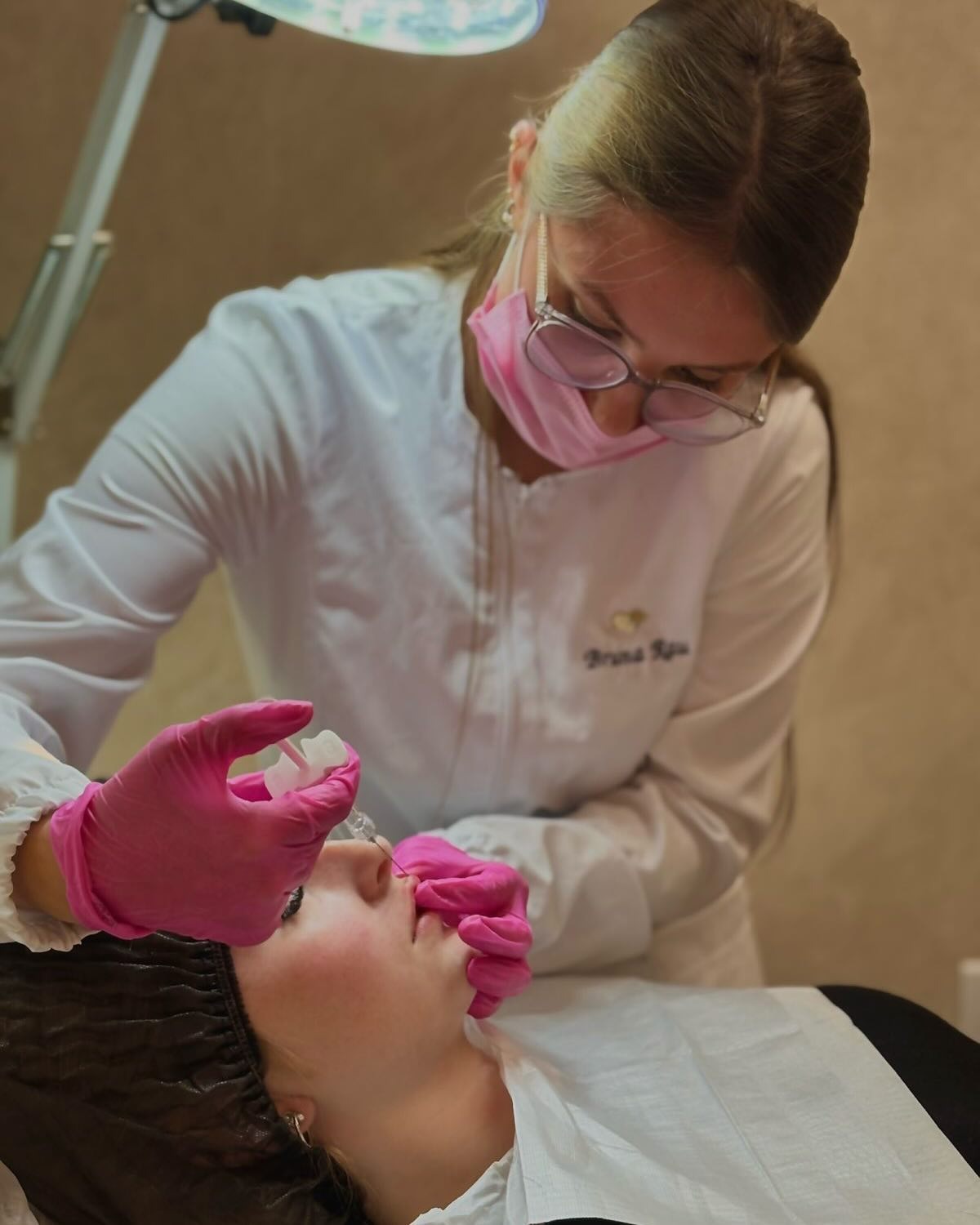 Dra. Bruna Rau atendendo uma paciente com máscara e luvas cor de rosa, realizando procedimento de estética facial